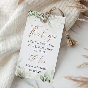 Könnte beinhalten: Ein weißes Geschenk-Etikett mit goldfarbenem Schriftzug "Thank you". Der Text "FOR CELEBRATING THIS SPECIAL DAY WITH US with love JOHN & SARAH 20.10.2026" ist ebenfalls enthalten. Es ist mit grünen und goldenen floralen Designs verziert und mit einer Schnur befestigt.