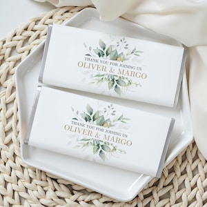 May include: Two chocolate bars with silver foil wrapping, resting on a white square plate. The bars feature a white label with a floral design and the text "THANK YOU FOR JOINING US OLIVER & MARGO" in gold lettering.