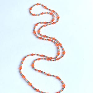 Könnte beinhalten: Eine lange Perlenkette mit orangefarbenen, roten, blauen und goldenen Perlen. Die Halskette besteht aus kleinen, runden Perlen, die zu einem zarten und farbenfrohen Accessoire aufgereiht sind. Die Perlen sind in einem sich wiederholenden Muster angeordnet.
