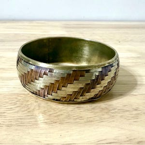 May include: A wide, woven metal bracelet with a mix of bronze, silver, and gold tones. The bracelet has a basket weave pattern and a smooth, polished interior. The bracelet is a piece of jewellery.