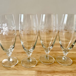May include: Four clear stemmed glasses with etched floral designs. The glasses have a tulip shape and sit on a round base. The glasses are arranged on a light brown wooden surface.