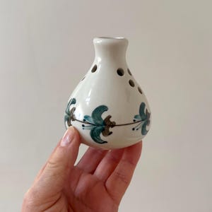 May include: A white ceramic bud vase with a bulbous body and a narrow neck. The vase features a floral design in teal, brown, and blue. It has several small holes around the body. The vase is held by a hand.