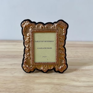 May include: A gold-plated frame with ornate detailing and a black velvet border. The frame is rectangular and features the text "TABLETOP MEMORIES" and "GOLDPLATED FRAME". The frame is 5.08 cm x 7.62 cm.