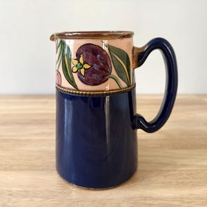 May include: A ceramic jug with a navy blue base and a light pink upper section. The upper section features a floral design with purple tulips and green leaves. The handle is navy blue, and the rim is brown.