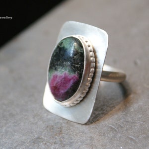 May include: A silver ring with a green and pink oval gemstone set in a square bezel. The ring has a beaded edge around the stone.