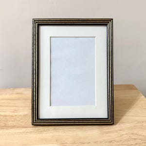 Op de afbeelding: Een rechthoekige fotolijst met een zwarte en gouden rand. De lijst heeft een witte passe-partout en ruimte voor een foto. De lijst staat op een houten oppervlak.