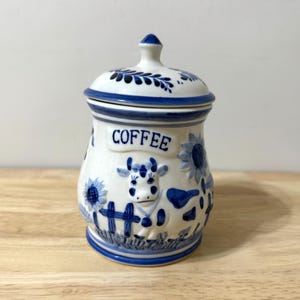 May include: White ceramic coffee jar with a blue and white cow and sunflower design. The word "COFFEE" is printed in blue. The lid and base have blue floral patterns.