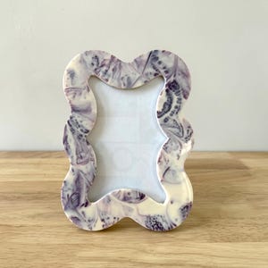 Peut inclure: Un cadre photo décoratif avec un bord ondulé unique. Le cadre présente un motif marbré dans des tons de violet et de blanc. Le cadre est fait d'un matériau brillant et est conçu pour contenir une photo rectangulaire.