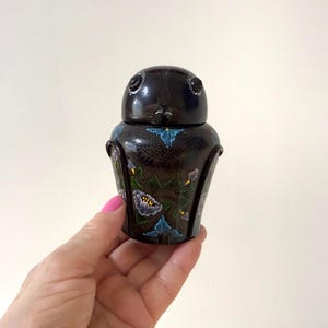 May include: A dark brown, rounded lidded container featuring a hand-painted floral design with blue, white, and green accents. The lid is smooth and rounded, while the body displays intricate patterns of flowers and leaves across its surface.