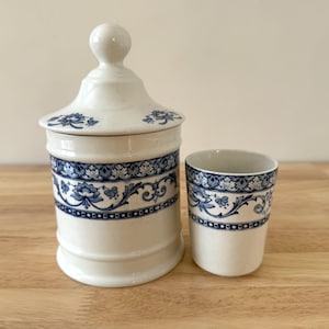 May include: A white ceramic jar with a lid and a small cup, both decorated with blue floral patterns. The jar has a rounded body and a knob on the lid. The cup is cylindrical. Both items are on a wooden surface.