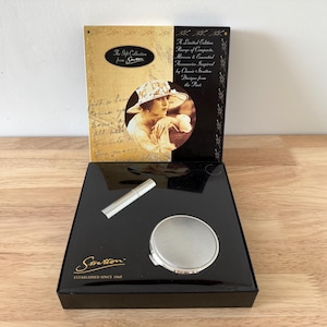 May include: A black box with a silver compact mirror, a silver lipstick tube, and a black and gold box with a vintage illustration of a woman in a hat. The box has the text "The Gift Collection from Stratton" and "Established Since 1960".