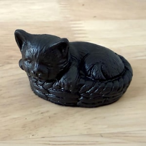 May include: A small, solid black figurine of a cat curled up and sleeping inside a woven basket. The cat has visible fur texture and pointed ears, while the basket features a detailed, interlaced pattern. The entire object is monochromatic black.
