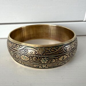 May include: A wide, brass-coloured bangle bracelet with intricate floral and leaf patterns. The bracelet has a slightly aged appearance, with some areas showing a darker patina. The design includes small, turquoise-coloured accents.