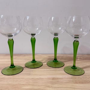 Op de afbeelding: Vier heldere wijnglazen met geëtste bloemmotieven. Elk glas heeft een levendige groene steel en voet. De glazen staan op een lichtgekleurd houten oppervlak, wat een aantrekkelijke presentatie creëert.