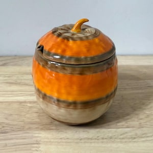 May include: A ceramic pumpkin-shaped jar with a lid and a small handle. The jar is painted with horizontal bands of orange, brown, and beige. The lid has a small, curved handle. The jar is sitting on a wooden surface.