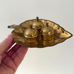 May include: A gold leaf-shaped tray with three small, round, gold containers with lids. The containers have a knob on top.