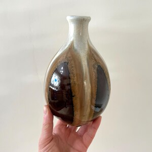 May include: A ceramic vase with a rounded body and a slender neck. The vase has a brown, tan, and cream colour scheme. The vase is approximately 18 cm tall and is suitable for displaying flowers or as a decorative item.
