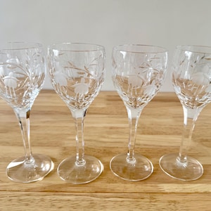 May include: Four clear crystal wine glasses with etched floral patterns. The glasses have a tulip shape, long stems, and round bases. The etched design features a floral pattern with delicate leaves and flowers. The glasses are arranged on a light brown wooden surface.