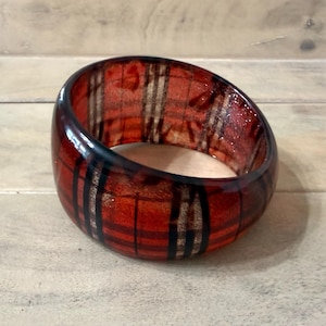 May include: A wide, circular bangle bracelet featuring a glossy, translucent finish with a prominent red and black plaid pattern. The design consists of intersecting horizontal and vertical lines that wrap around the entire exterior surface of the band.