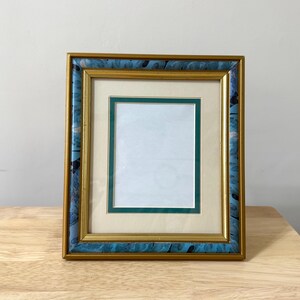 May include: Square picture frame with a gold inner frame and a teal and gold outer frame. The frame has a cream-coloured mount with a green border. The frame is on a wooden surface.
