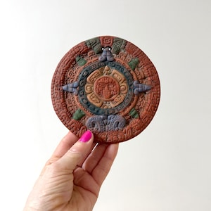 Op de afbeelding: Een ronde Azteekse kalender in terracotta tinten met ingewikkelde gravures. Het centrale ontwerp toont een zonachtig gezicht, omringd door concentrische cirkels in bruin, blauw en groen. De kalender wordt vastgehouden door een hand met roze nagellak.