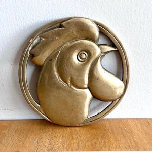 May include: A gold-toned metal relief of a rooster's head, presented in profile within a circular border. The rooster's features, including its comb and beak, are well-defined. The piece has an aged appearance, possibly an antique decorative item.