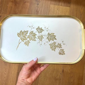 May include: An oblong serving tray with a white surface and a gold-toned metal rim and handles. The centre of the tray features a decorative botanical illustration of gold-coloured maple leaves and delicate vine tendrils across the white base.