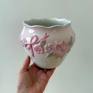 Op de afbeelding: Een lichtroze keramische bloempot met een geschulpte rand. De bloempot is versierd met in reliëf aangebrachte roze bloemen, een strik en groene bladeren. Het bloemmotief loopt rond de buitenkant van de bloempot.