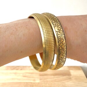 May include: Two gold-colored bangles are worn on a wrist. One bangle has a textured, scale-like pattern, while the other features an etched floral design. The bangles are displayed on a light wooden surface.