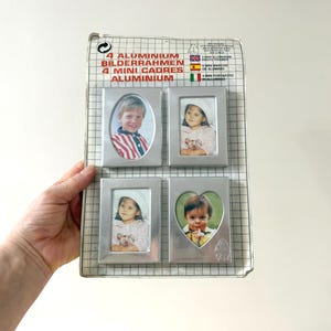Op de afbeelding: Een set van vier kleine, zilverkleurige aluminium fotolijsten in verschillende vormen: ovaal, rechthoekig en hartvormig. De lijsten bevatten foto's van kinderen. De verpakking leest "4 ALUMINIUM BILDERRAHMEN 4 MINI CADRES ALUMINIUM."