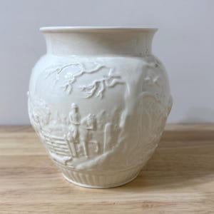 May include: A white ceramic vase with a detailed relief design. The vase features a rounded shape with a wide opening and a detailed scene of figures and trees. The relief is in a classic style, adding a touch of elegance to the decorative piece.