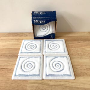 May include: A set of six square ceramic coasters featuring a white surface with a blue spiral pattern. The coasters are presented alongside their original box, which displays the brand name "Pilkington's". Each coaster measures approximately 10 cm x 10 cm.