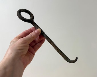 May include: A vintage, dark metal hook with a circular handle and a curved end. The hook is held in a hand against a plain white background. The metal has a weathered, aged appearance.
