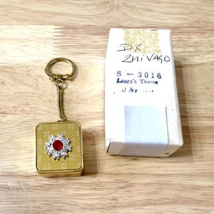 May include: A gold-coloured rectangular keyring with a red and clear rhinestone flower design. The keyring is attached to a gold-coloured ring and a coiled spring. A white box with handwritten text is next to the keyring.