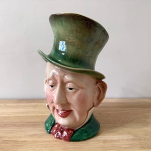 May include: A ceramic figurine of a man's head, featuring a green top hat and a green jacket. The face is detailed with rosy cheeks, a red bow tie, and a slight smile. The figurine is set on a wooden surface.