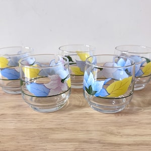 May include: Five clear, round drinking glasses with a floral design. Each glass features a band of blue, yellow, and pink flowers with green accents. The glasses are arranged on a light brown wooden surface.