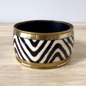 May include: A round bracelet with a black interior and a brass-colored rim. The exterior features a zebra stripe pattern in black and white. The bracelet is sitting on a light-colored wooden surface.