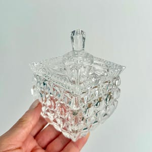 May include: A clear, square-shaped crystal jar with a lid, held by a hand. The jar features a diamond-cut pattern, creating a sparkling effect. The lid has a small, faceted handle. The overall design is elegant and decorative.