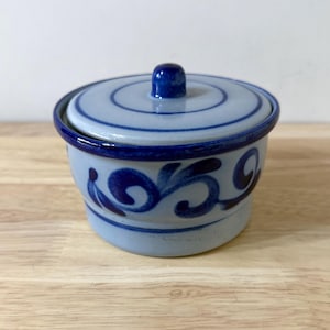 May include: A light blue ceramic lidded jar featuring a dark blue hand-painted floral scroll pattern around the exterior. The lid has a matching dark blue knob handle and concentric circular rings, with a dark blue rim around the base and top.