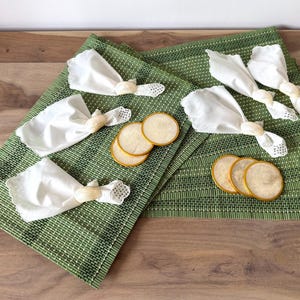 May include: A table setting with green placemats, white napkins held by decorative napkin rings, and gold-edged coasters. The placemats have a woven texture, and the napkins are folded in a decorative style.