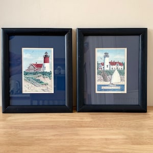 May include: Two framed prints showcasing coastal scenes. Each print features a lighthouse with a red roof and white tower, set against a blue sky. One print includes a sailboat. The frames are black, and the prints are matted in navy blue.