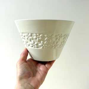 May include: Cream-coloured ceramic bowl with a flared shape. The bowl has a band of embossed floral designs around the middle. The bowl is held in a hand against a white background. The bowl is likely used for serving or display.