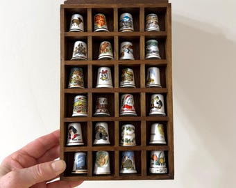 May include: A wooden display case filled with various decorative thimbles. The thimbles are white with colorful designs, and the case has a dark brown finish. The thimbles feature images of landmarks, characters, and holiday themes.