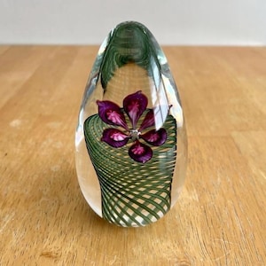 May include: A clear glass paperweight with a purple flower design. The flower has a crystal center and is surrounded by green, woven glass. The paperweight is egg-shaped and sits on a wooden surface.