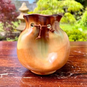 May include: A small, brown and green ceramic vase with a unique, rounded shape and a leather cord tied around the opening.
