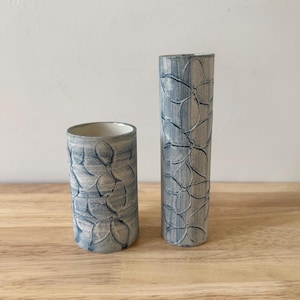 May include: Two ceramic vases with a floral pattern. The vases are cylindrical, one short and wide, the other tall and slender. The floral design is embossed and painted in shades of blue and gray, creating a textured appearance. The vases are displayed on a wooden surface.