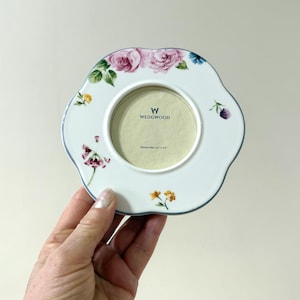 May include: White ceramic picture frame with a scalloped edge, adorned with floral designs in pink, purple, yellow, and green. The frame has a blue trim and features the "W WEDGWOOD" logo. Picture size is 3.5" x 3.5".
