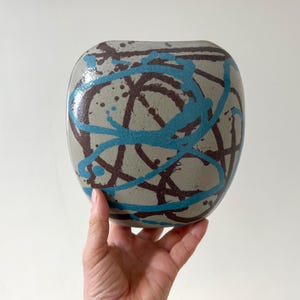 May include: A ceramic vase with a rounded shape, featuring a grey base with brown and teal abstract paint strokes. The vase is held by a hand against a white background. The design gives the vase a modern, artistic look.