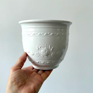 May include: White ceramic planter with embossed floral and geometric designs. The planter has a rounded shape with a slightly flared rim. The design includes flowers, leaves, and geometric patterns. The planter is held in a hand.