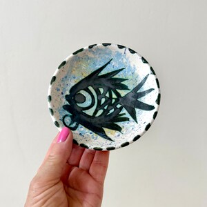 May include: A small round ceramic dish featuring a hand-painted black fish design in the center. The background of the dish is speckled with light blue and yellow paint, and the outer rim is decorated with a series of small black dots.